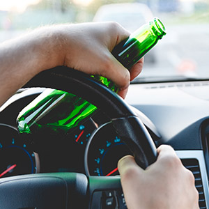 Driver holding a steering wheel with an open beer bottle, illustrating DWI or drunk driving charges and criminal law.